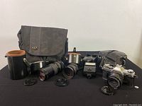 Full view of Canon AT-1 camera, two lenses (80-200mm zoom and 28mm prime), Canon Speedlite 155A flash, camera bag, and lens cases arranged on table.
