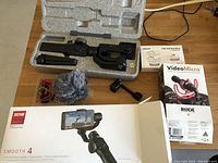 Zhiyun Smooth 4 gimbal in foam case, Rode VideoMicro microphone with red shock mount and deadcat windscreen, triple cold shoe mount and instruction manual all arranged on wooden floor.