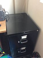 Top and front angle of the black filing cabinet showing two drawers, handles, and label holders. Surface has slight wear.
