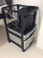 Front angled view of black plastic office cabinet showing two light grey drawers, black top bin, metal wire racks on sides, and caster wheels.
