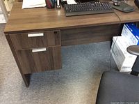 Front and left side view of the dark brown desk, showing two drawers with silver handles and the cable passthrough hole on the top surface.
