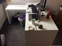 White L-shaped desk setup with computer equipment, tape, measuring tape, pen, and mug on top. Smaller side desk surface in line with main desk.