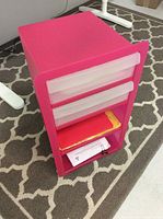 Front angle view of pink plastic office cabinet showing two translucent drawers and open shelves with some documents inside.