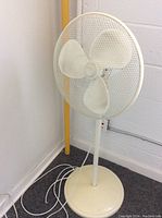 Front view of white plastic standing fan with three blades and mesh cage. Fan is plugged in with visible power cord coiled on the ground.