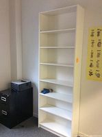 Tall white bookshelf with seven shelves placed against a wall near a black filing cabinet and printer, showing its full height and width.
