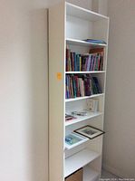 Tall white wooden bookcase with seven shelves, positioned against a wall with some books and papers placed on three of the shelves.