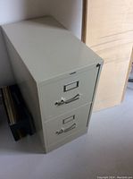 Photo of one beige two-drawer filing cabinet standing on floor, showing front and side view with closed drawers