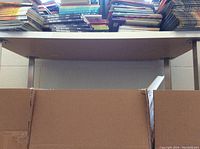 Photo showing metal legs and underside of white rectangular desk top beneath stacked books and cardboard boxes.