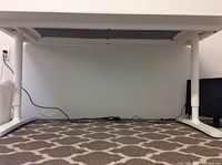 View under the desk showing white legs and power strip on carpeted floor with patterned area rug.