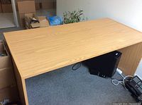 Wide wood laminate desk with flat edges and plain design, positioned on carpeted floor near wall, with some boxes and cables underneath.