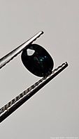 Close-up photo of an oval cut blue-green sapphire gemstone held in tweezers showing its color and cut facets.