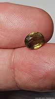 Close-up of the oval green yellow sapphire gemstone held between fingers showing clarity, color variation, and natural inclusions.
