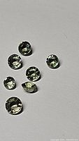 Seven round cut moss green sapphires with visible mossy inclusions and good luster arranged on a light background.
