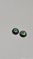 Close-up photo of two faceted round sapphires showing their multicolored teal, green, and blue hues on a plain background.
