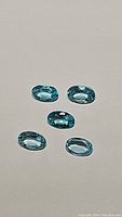 Five oval-shaped natural blue apatite gemstones with bright paraiba blue coloration placed on a neutral background.