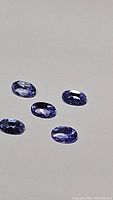 Five faceted oval cut purple blue tanzanite gemstones arranged on a white background, showing color and clarity.