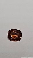 Photo showing a cushion cut natural African zircon with warm brownish-orange tone, moderate internal clarity, lying on white background.