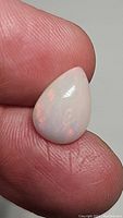Close-up photo of pear-shaped fire opal cabochon held between fingers showing its color and surface details.
