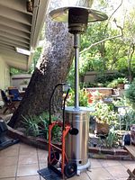 Outdoor setting image showing tall propane gas patio heater with reflector top and adjacent orange and black moving dolly under a covered patio.