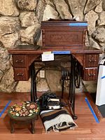 Front view of the full sewing machine setup including wooden table with drawers, foot pedal, round floral footstool, and woven throw on floor.