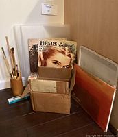 Full view of art tools lot arranged on floor showing holding bamboo container with paint brushes, books including 'Heads from Life', translucent papers and art folders/boards, and a cardboard box with supplies.
