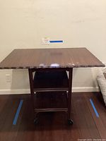 Front view of dark wood table showing scalloped edged top, fold-down ends, two lower shelves, and casters.