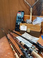 Fishing rods with reels laid on table next to tackle box and a boxed vertical rod rack, fish net leaning against wood paneled wall.