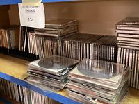 Shelf with flat black CD holder rack holding classical music CDs, plus stacks of CDs horizontally placed on a wooden shelving unit.