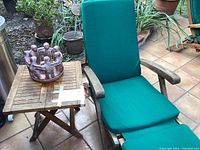 Teak lounge chair with green cushion next to teak side table on tiled floor, ceramic candle holder on table, outdoor patio setting with plants.