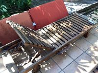 Teak outdoor lounge chair showing the adjustable slatted backrest and orange cushion behind it, placed on tiled patio under dappled sunlight.