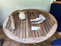 Round teak table top with slatted surface and four decorative art pieces: driftwood branch, carved face, wrought iron holder, driftwood antler