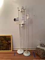 Two white floor lamps standing side by side against a wall, each with three cylindrical spotlight heads arranged on vertical poles with round bases.