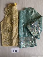 Two sleeveless vintage tops, one gold with floral bead and sequin embellishments and one light blue-green with dense sequin coverage and floral patterns, laid flat on a surface.