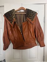 Brown leather bomber jacket hanging on wooden hanger showing front and leopard print collar details.