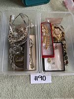 Two plastic bins containing silver-colored tiaras, rhinestone jewelry, wristwatches with metal bands, and other costume jewelry pieces.