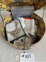 Photo of decorative round hat box with costume jewelry inside, including bib necklace in display box, rhinestone bracelet, and several small bags with additional earrings and items.