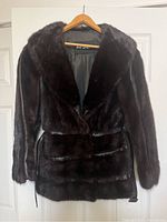 Front view of dark brown Holt Renfrew vintage fur coat on hanger against white door background. Shows plush fur texture, collar, front pockets, and belt detail.