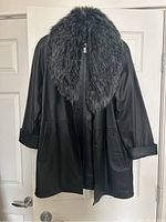 Front view of black leather jacket with large faux fur collar hanging against door.