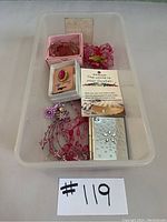 Plastic bin containing multiple pink costume jewelry items including earrings, brooches, necklace, and pins, arranged with price tag #119.