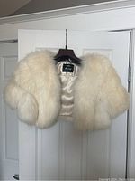 Front view of a white fur bolero jacket hanging on a wooden hanger against a white door.