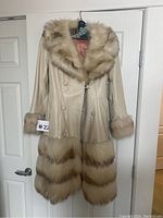 Full view of the vintage leather coat with fur trim hanging on a door, showing cream leather and fur at collar, sleeves, and hem.