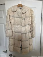 Front view of the vintage long white fur coat hanging on a wooden hanger against a white door. The fur is plush and arranged in horizontal panels with a wide collar.
