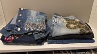 Folded denim clothing including a blue denim jacket with bead embroidery and two pairs of embellished denim pants.