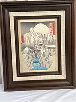 Framed Asian mountain landscape print showing house, river, rocky cliffs, and trees in muted colors with a tall wooden frame and fabric matting.