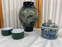 Full view of ceramic vase, blue and white covered jar, and two green ramekins on white surface against curtain background.