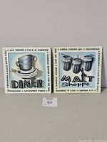 Two square 3D metal wall art pieces side by side on a gray wall, featuring diner and malt shop themes.