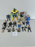 Lot of assorted action figures including a large green and yellow armored figure, small armored figures, and humanoid figures, posed and grouped on a white surface with a background.