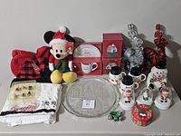Lot of Christmas-themed items displayed on a table including Mickey Mouse plush, red plaid textiles, glass serving platter, Peanuts mugs, ceramic snowmen, and small figurines