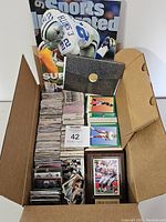 Box showing collection of football cards, a Sports Illustrated magazine, a black card holder, and a framed plaque