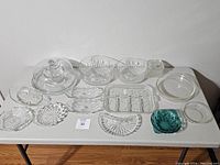 Wide view of multiple clear glass dishes, bowls, and plates with decorative pressed and cut glass details, arranged on a white table.